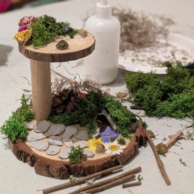 A craft fairy house in mid-construction, made out of wood, moss, stones, and flowers, on a white table next to a bottle of glue. 