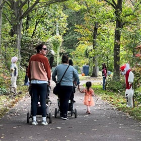 A group of woman walking with strollers on a paved path in a green outdoor setting with scarecrows lining the path. 