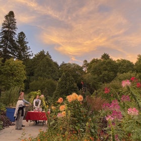 A table with wine and cheese is set up in a beautiful blooming garden as the sun sets and people mingle. 