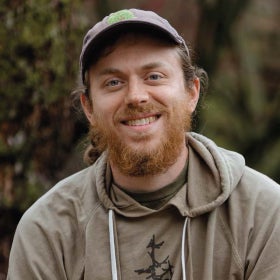 A headshot of a man with a baseball cap with an embroidery of a tree and a red beard, wearing a khaki sweatshirt.
