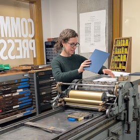 A woman in glasses stands at a printing press holding up a plate. 
