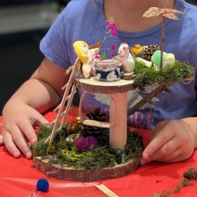 A child's hand surround a tiny structure made of wood and decorated with whimsical items