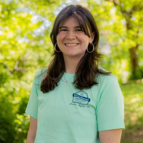 A headshot of a young woman with gold hoops and a septum piercing wearing a pastel green shirt outdoors. 