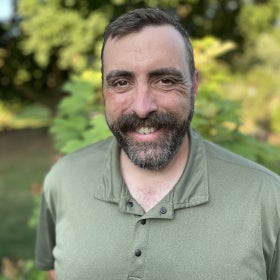 A head shot of a smiling man with a beard wearing a green collared shirt outdoors. 
