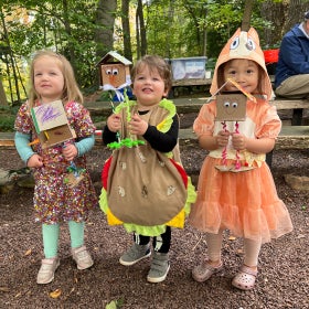 Three small children in Halloween costumes stand outdoors in a wooded area holding a craft they made. 
