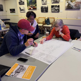 Three people sit at a table looking at large plans on a white table. 