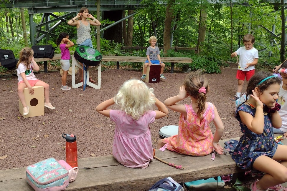 A group of young children sit in an outdoor classroom with their hands to their ears looking at the teacher. 