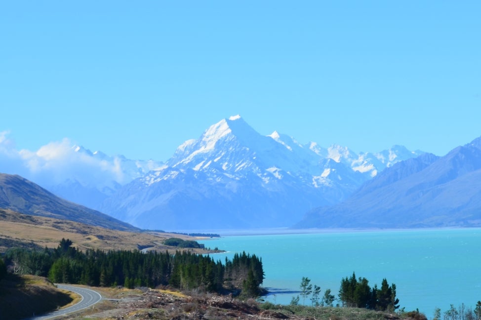 A mountain view of Mt. Cook, New Zealand with a road and trees, as well as a blue body of water, in the foreground. 