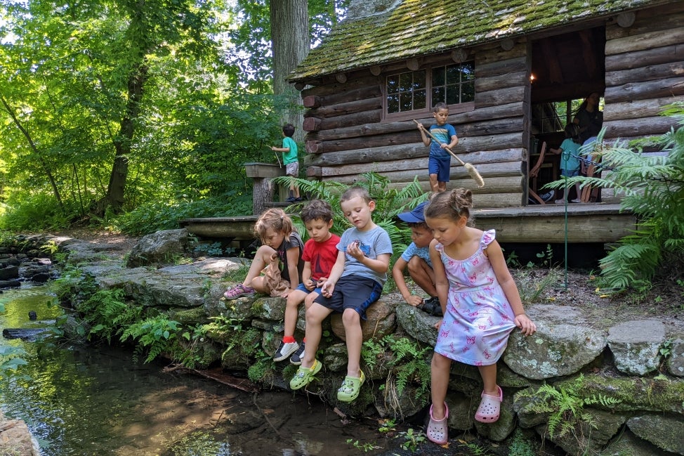 A group of five young children sit on a rock ledge on the side of creek with a log cabin in the background. 