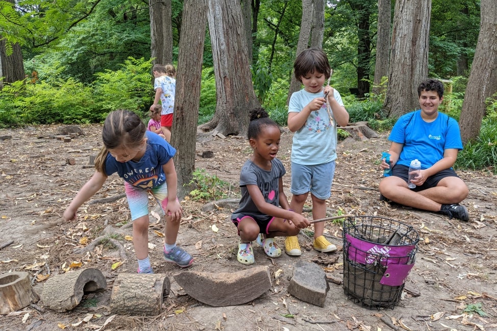 A group of young children build small houses out of natural materials in the woods. 