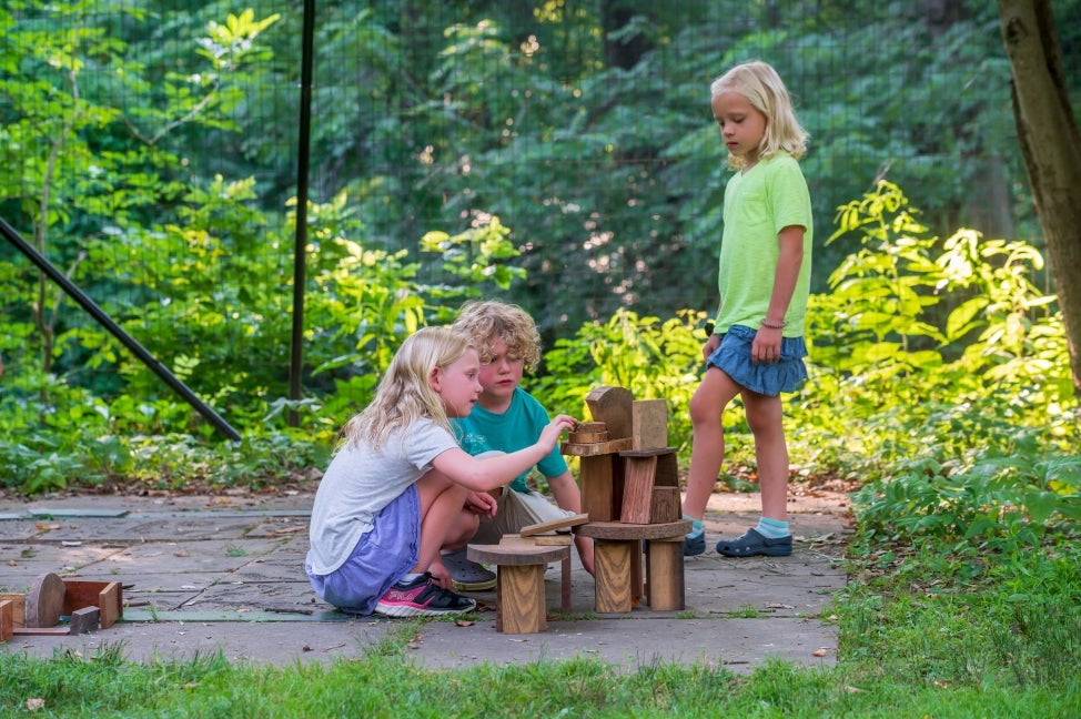 Three children with blond hair build a small fairy house out of wood blocks outdoors.
