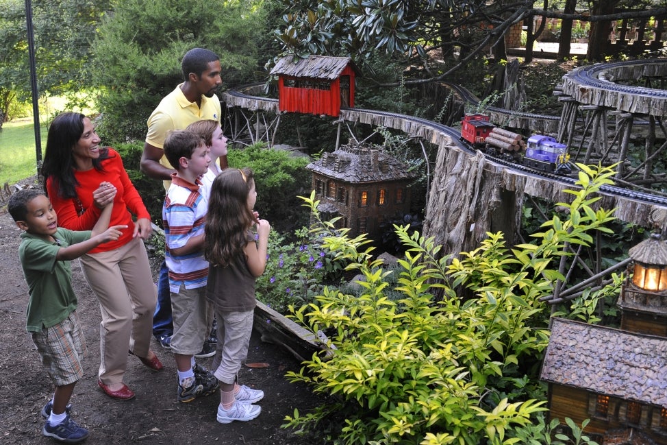 A family watched a model train on an outdoor track surrounded by plants and miniature buildings. 