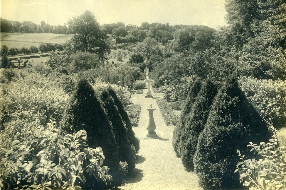 A black and white photograph of a garden with a sundial and fountain. 