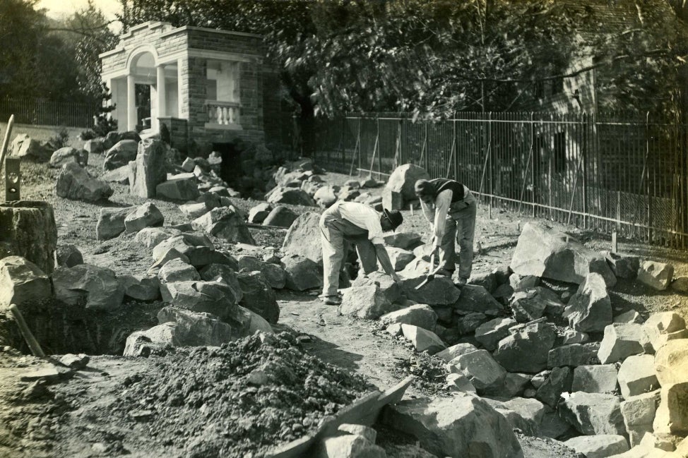 A black and white photo of two men lifting large boulders in a garden.