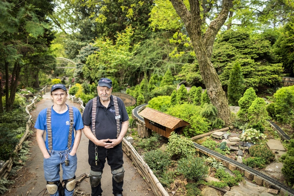 A young man and an older man wearing hats and thick suspenders stand outdoors within a model train display.