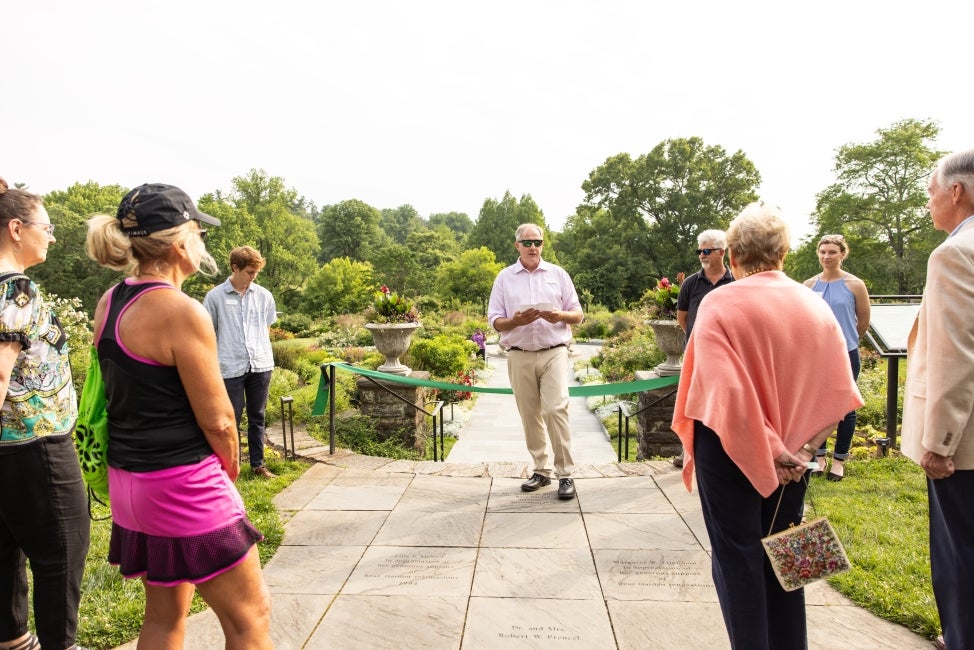 A man in sunglasses stands in front of a ribbon and addresses a group of people in a public garden.