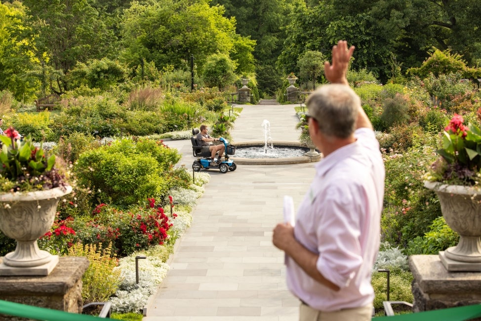 A man waves towards a person riding a mobility scooter in a public garden.