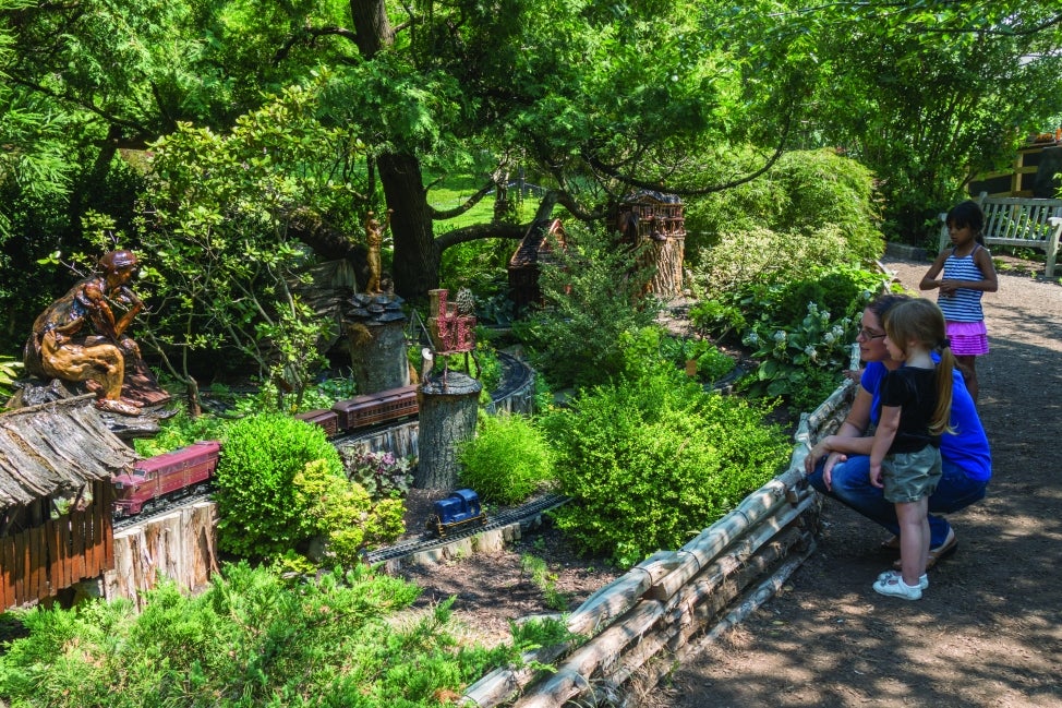 A woman and a young girl smile at a miniature train display in a public garden.