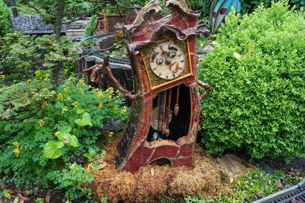 A small, fabricated grandfather clock with a human-like face surrounded by flowers and foliage.