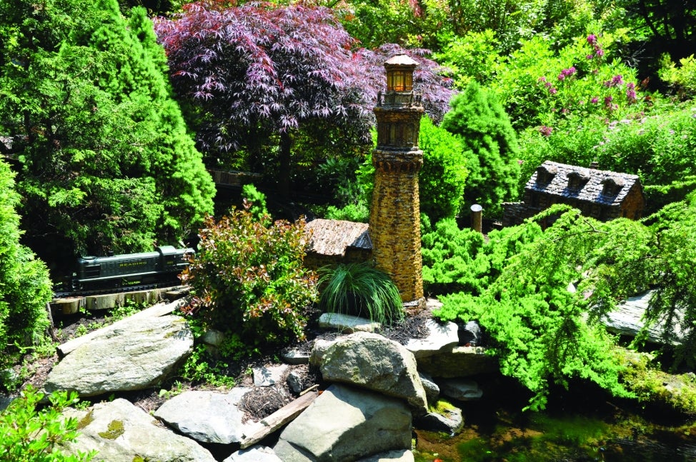 A miniature fabricated lighthouse set in a model train display surrounded by plants.