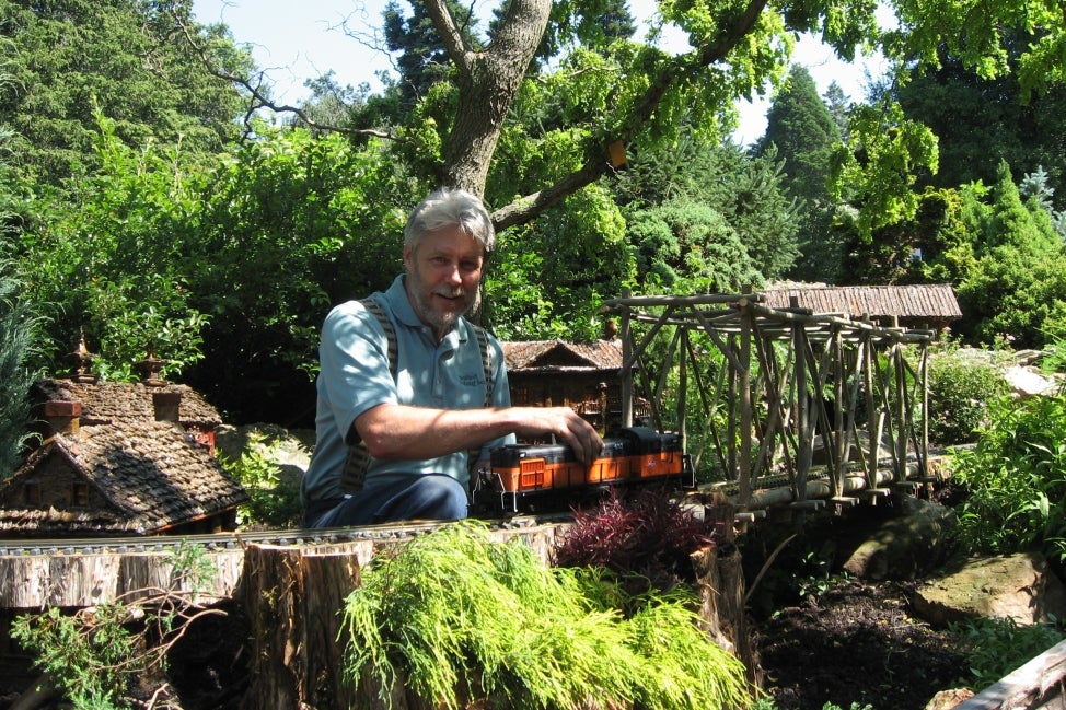 A man wearing suspenders hold a miniature train in an outdoor model train display.