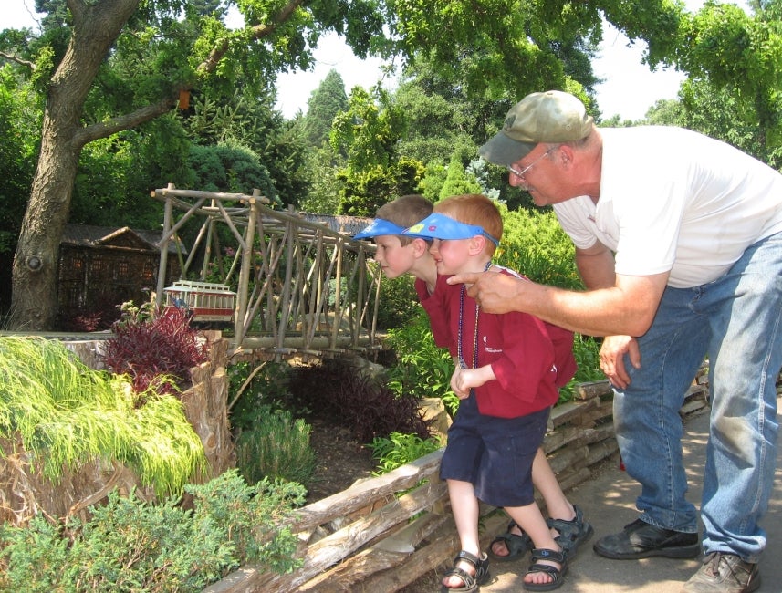 A an older and two young boys look at an outdoor model train display. 