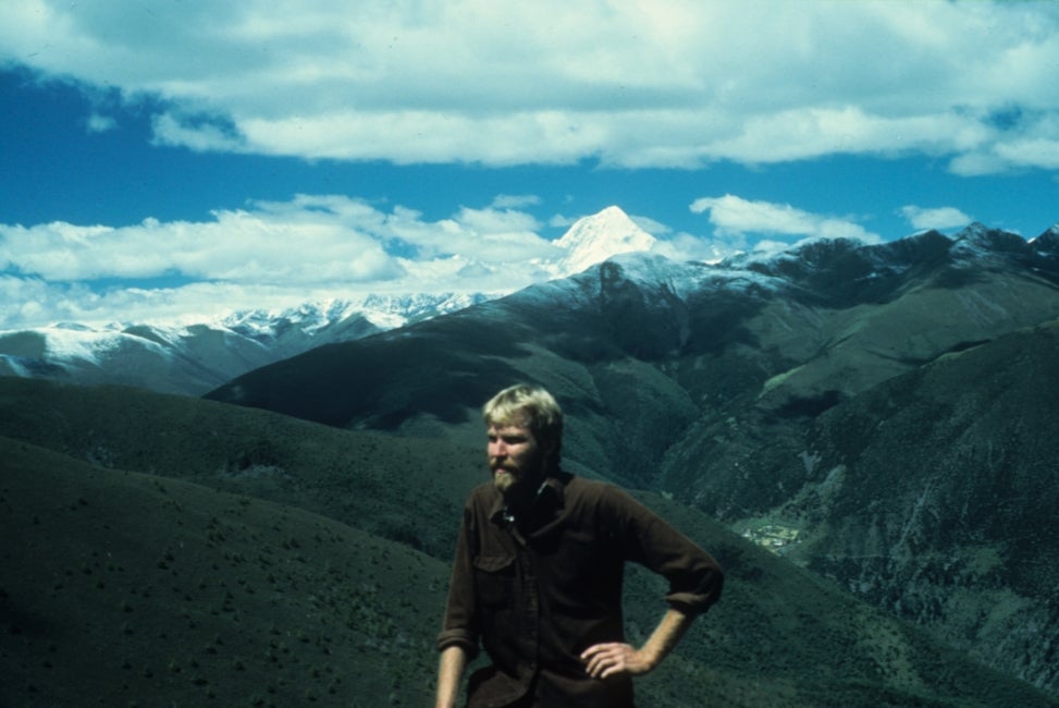 A man stands in the mountains with his hand on his hip and a blue sky in the background. 