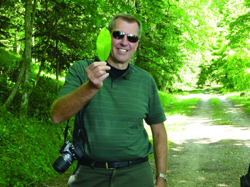 A man wearing green with a camera slung over his shoulder smiles and holds up a leaf. 