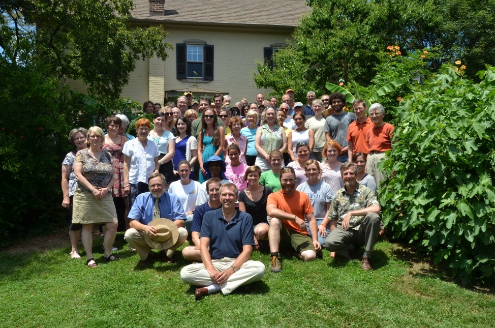 A large group of people pose for a staff photo in front a building and large green trees. 