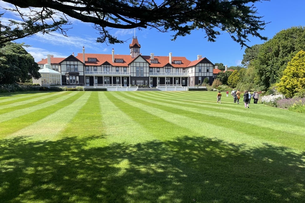 A large, ornate New Zealand government house in the background with a group of people walking towards it on a large lawn in the foreground on a sunny day. 