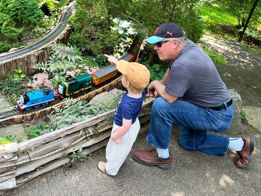 A grandfather and grandson watch a miniature train display in a public garden.