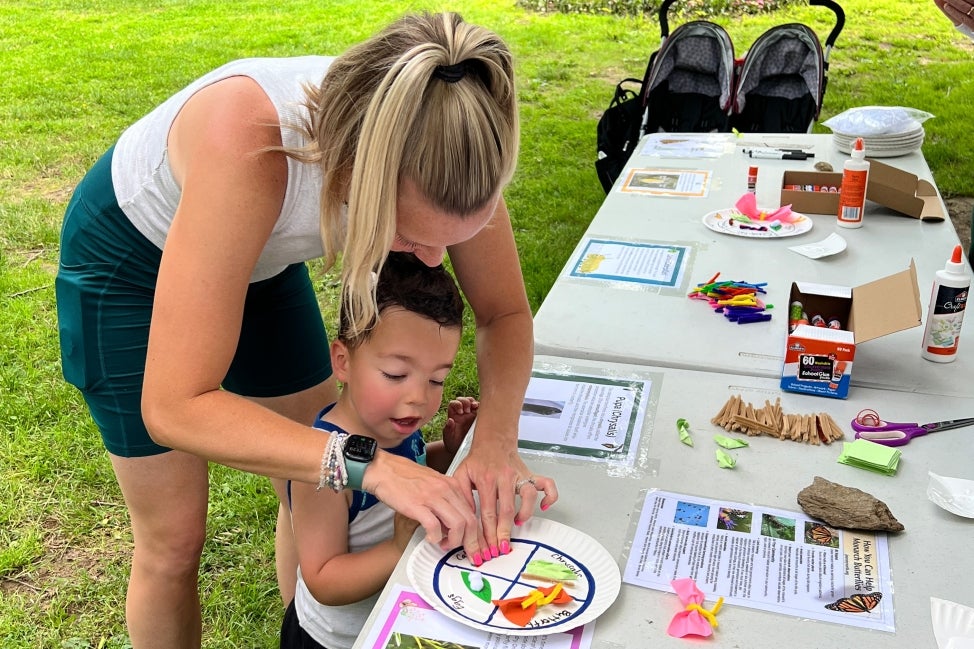 A mother helps her son with arts and crafts at a table outdoors in a public garden.