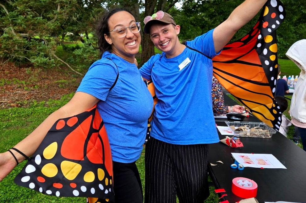 Two people in blue shirts and black pants wear a monarch butterfly wings costume outdoors smiling and posing for the camera. 