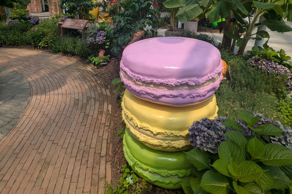 A large fabricated stack of three macarons in pink, yellow, and green set within a botanical conservatory and surrounded by plants. 
