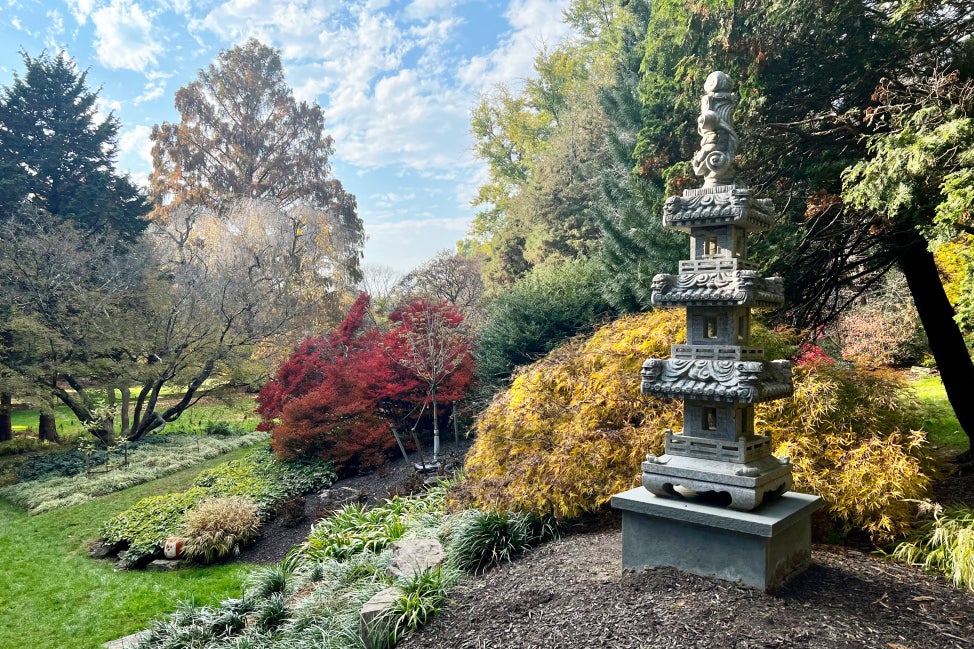 A Japanese-style garden with a concrete pagoda statue in the foreground and fall foliage in the background on a sunny day.