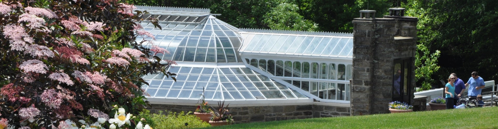 A glass greenhouse with a stone entry way with flowers on the left foreground and two people walking to the right.