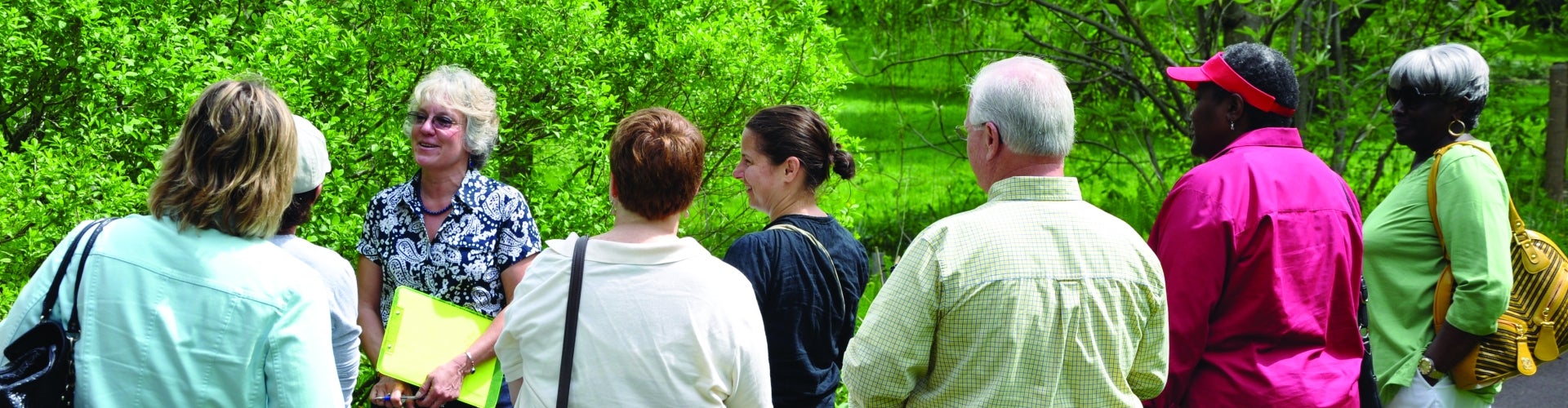 A group of people on a garden tour with bright green trees in the background. 