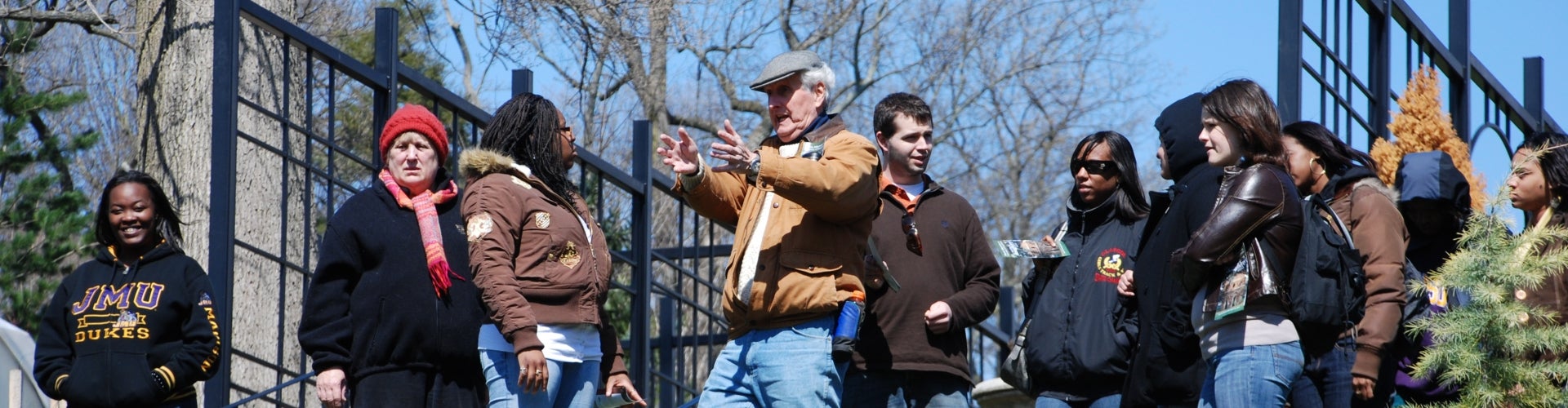 A group of ten people in winter coats are led on a tour of a public garden.