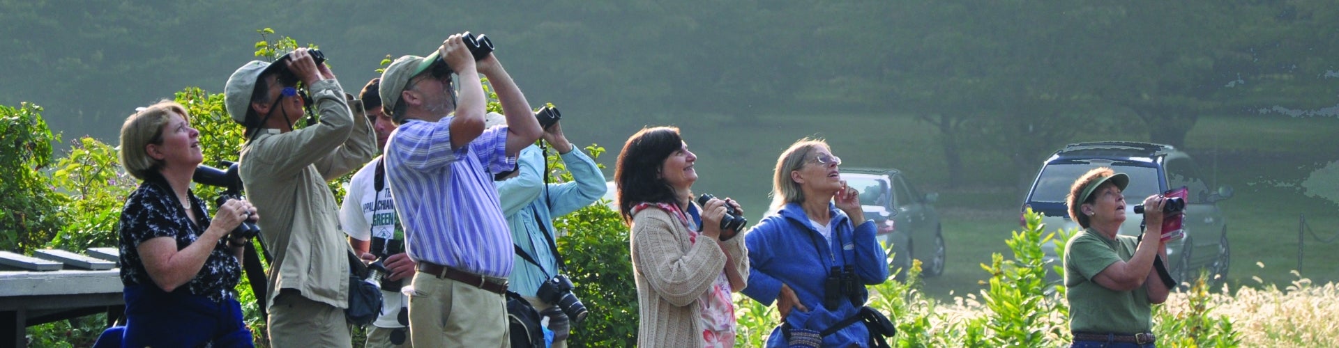 A group of people holding binoculars look up at the sky. 
