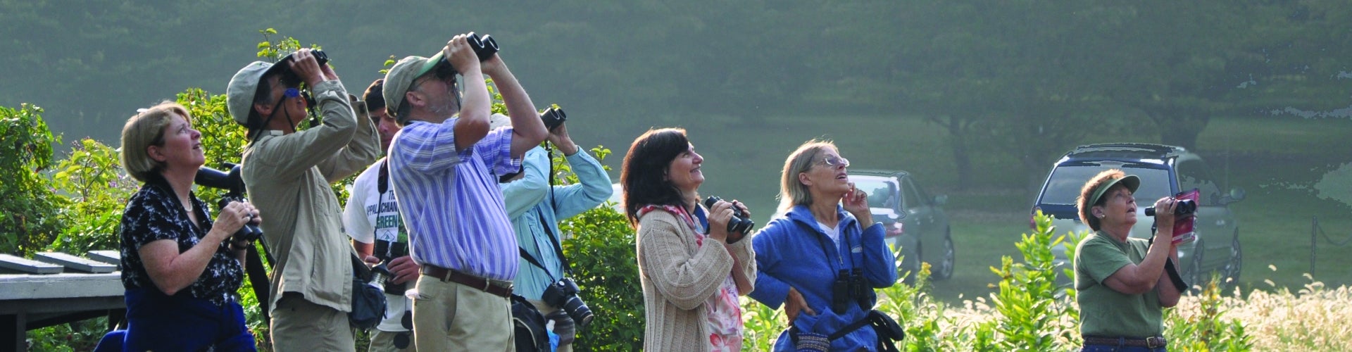 A group of people holding binoculars look up at the sky.