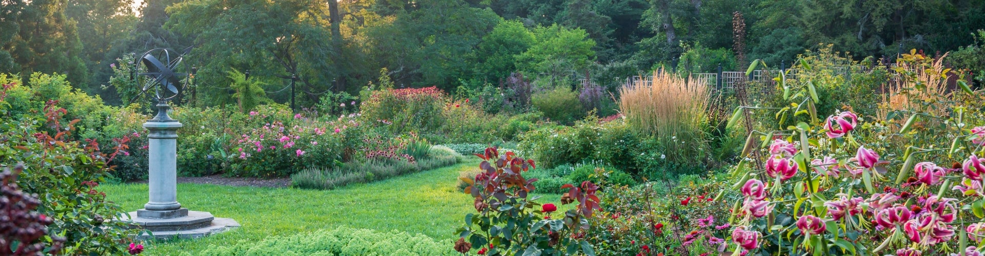 A stone and metal sculpture stands to the side of a colorful, blooming rose garden. 