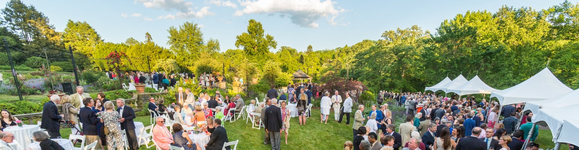 A large group of people mingle outdoors in a rose garden with tables, chairs, and tents.