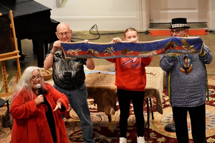 A woman in red holds a microphone while behind her stands three people holding up a long narrow tapestry of Lenape embroidery.