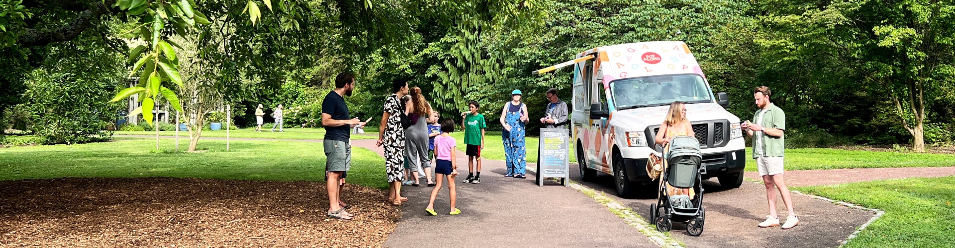 A group of people stand outdoors in a public garden in front of an ice cream truck eating ice cream on a sunny day.