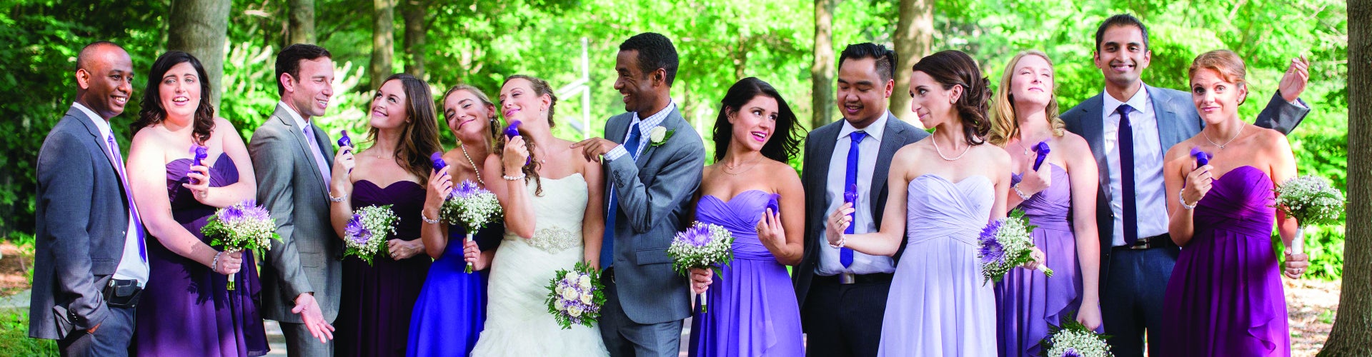 A bridal party dressed in purple and blue with the bride and groom stand in an outdoor green space laughing and having a good time. 