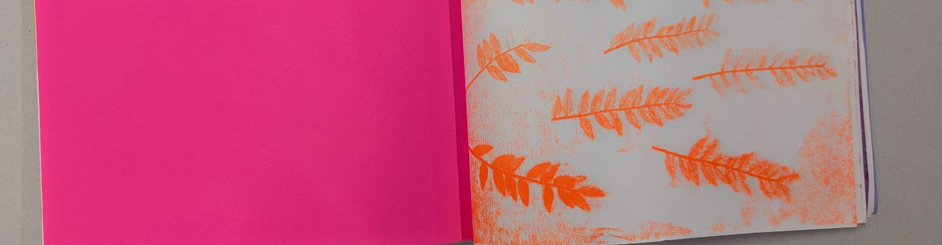 A hand-made book with a bright pink cover opened to the first page with orange prints of fern fronds on white paper. 