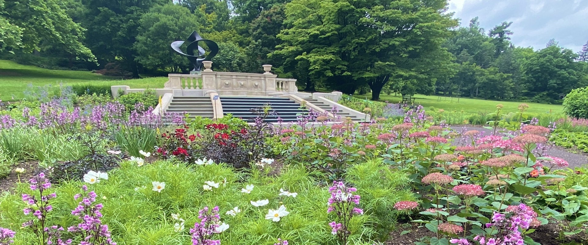 A metal sculpture sits atop a step fountain surrounded by flowers.