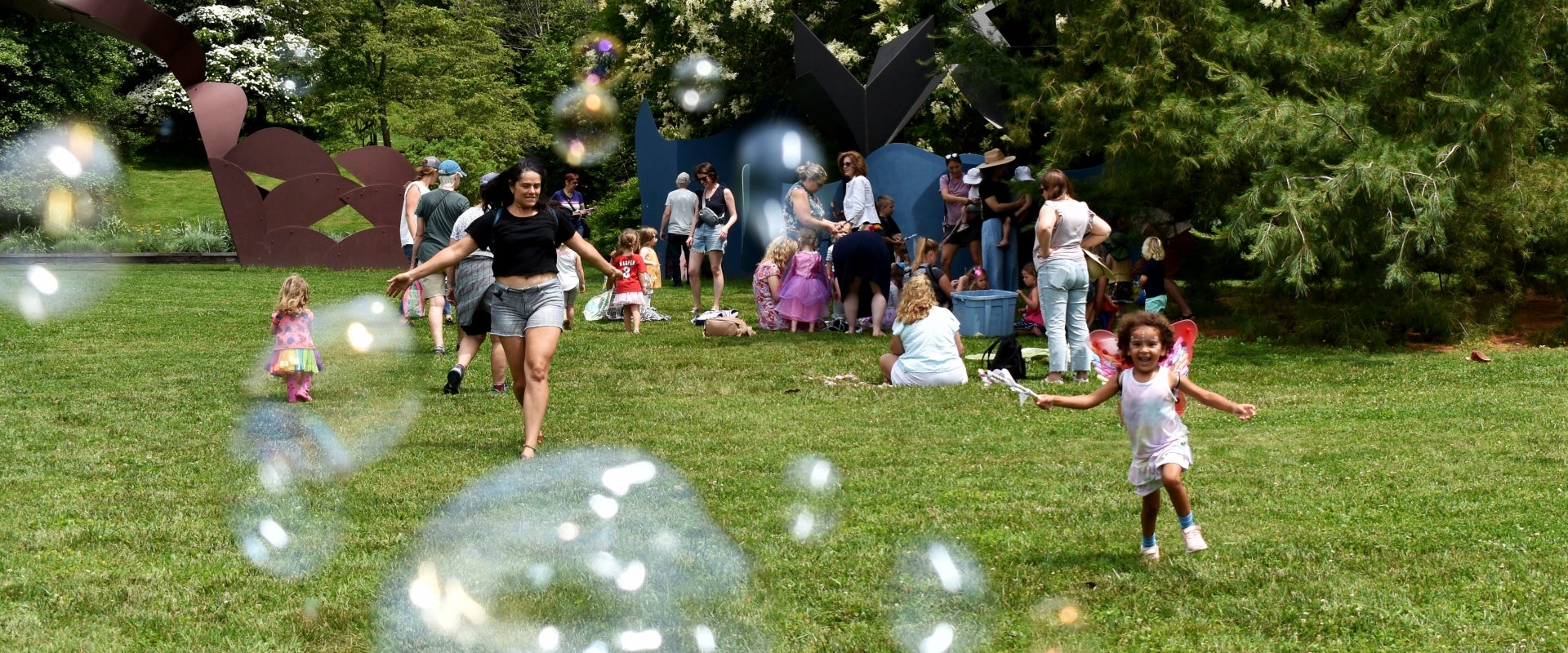 A group of people gathered on a lawn filled with bubbles as a mother and daughter run smiling. 