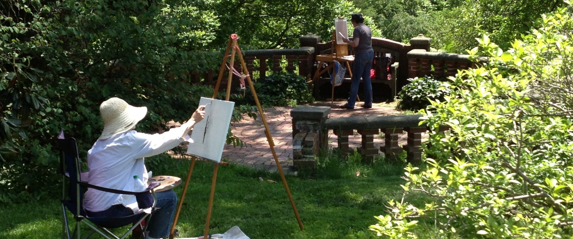 A person in a hat sits in a garden and paints at an easel. 