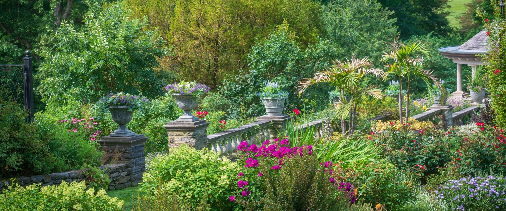 A garden in bloom with a stone wall decorated with planters and a gazebo. 